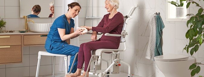 En kvinna som hjälper en dam med att tvätta händerna i ett badrum, damen sitter i en hygienstol från leverantör Etac.