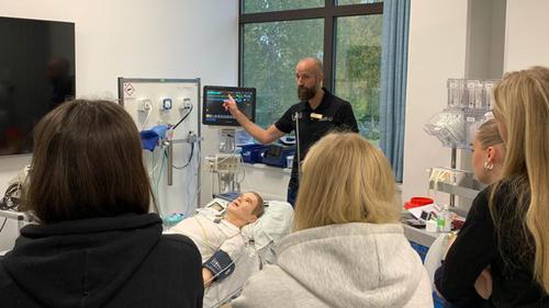 En utbildningsledare på Kliniskt träningscentrum visar och berättar om hur ett hjärtstopp behandlas. På en sjukhussäng framför honom ligger en övningsdocka och några elever på studiebesök står intill och lyssnar. 