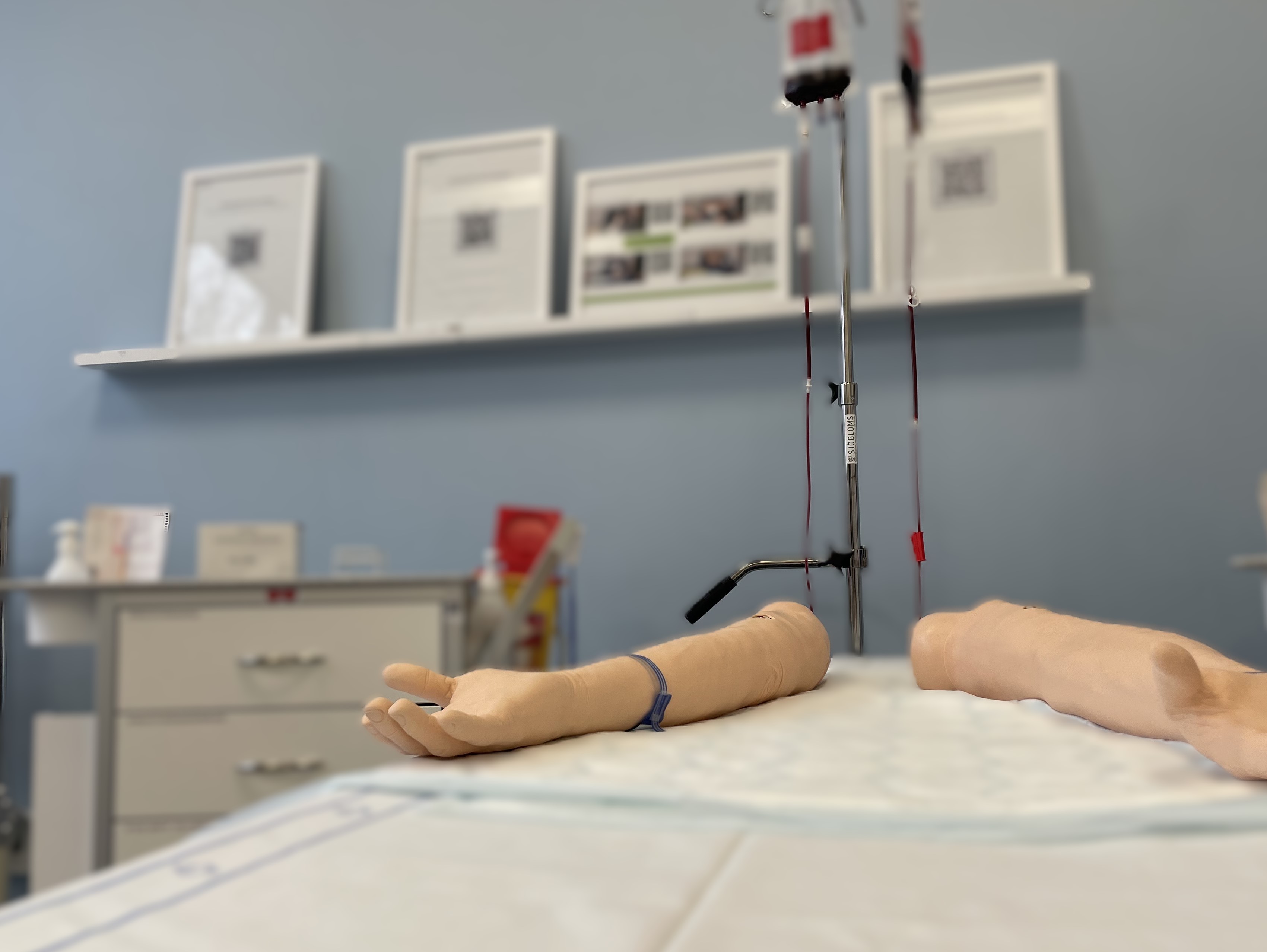 En bild på provtagningsarmar som används vid exempelvis övning av venös provtagning. I bakgrunden ses en provtagningsvagn och infusionspåsar med rött innehåll.