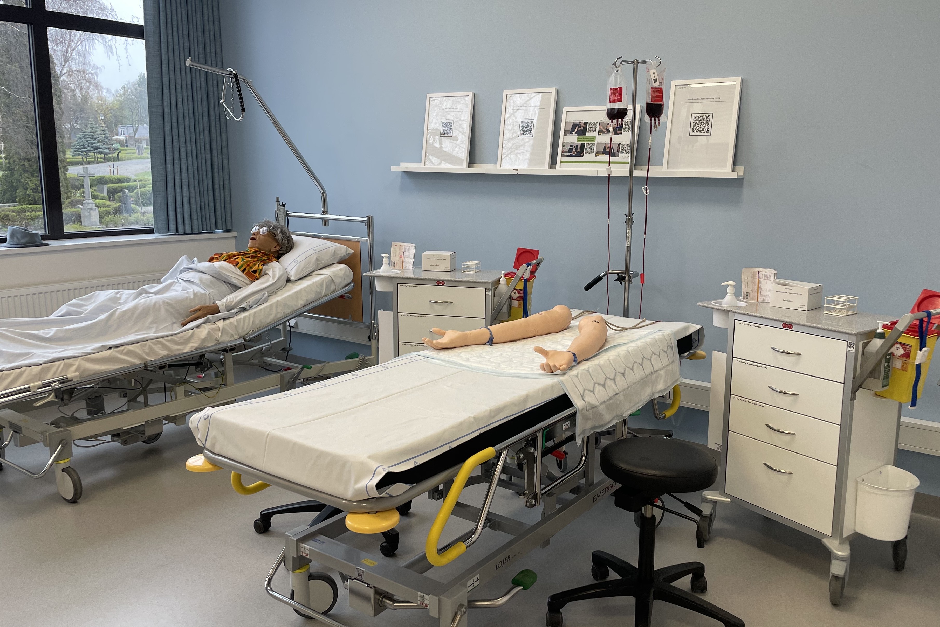 En bild på ett metodrum med delar av den utrustning som används vid färdighetsträning kompetenskort hela undersköterska. Du ser provtagningsarmar och vagn samt en simulatordocka som används vid genomgång av vitalparametrar.