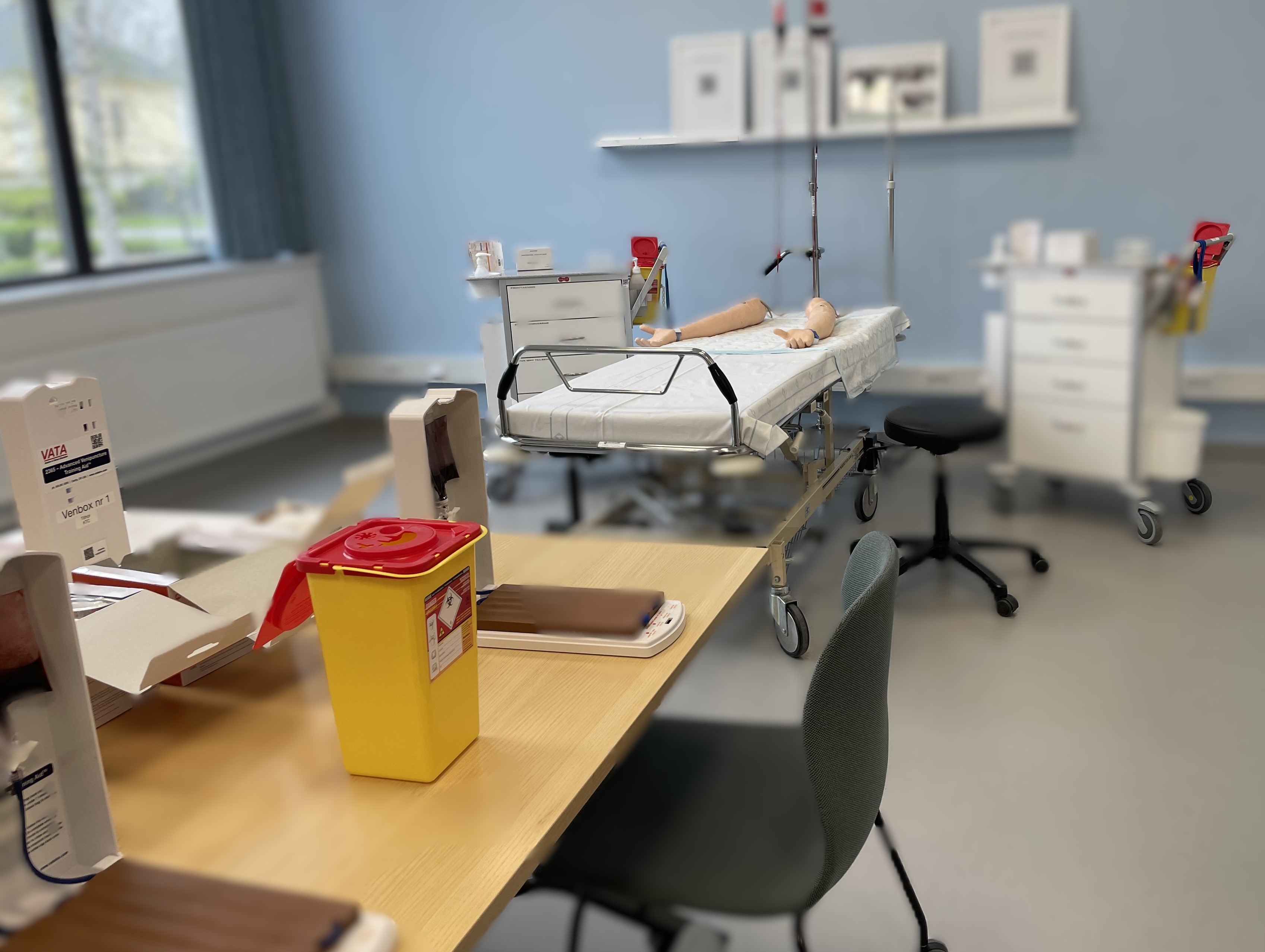En bild på ett metodrum med utrustning som används vid utbildningen perifer venkateter. Ett bord med venboxar där mängdträning sker och i bakgrunden ses provtagingsarmar där hela momentet genomförs.