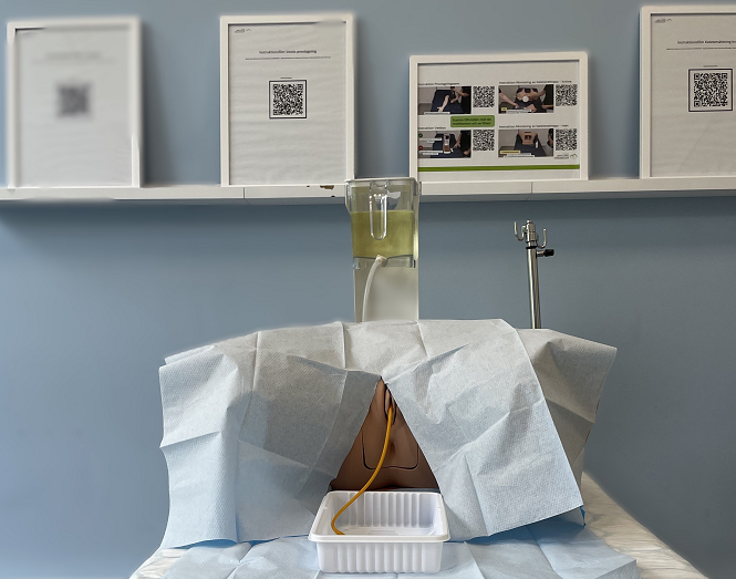 Bild på en kateterattrapp på en brits som används vid övning och examination av katetersättning. Attrappen har en kateter satt och slangen ligger i en skål som ligger på britsen.