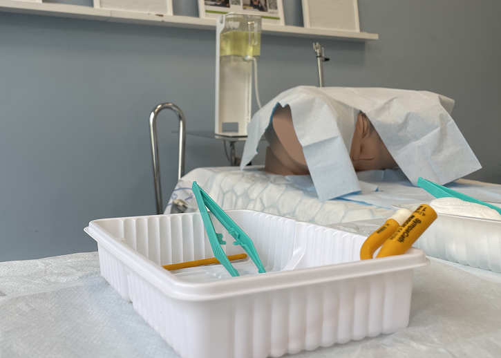 En bild på en inklädd kateterattrapp som ligger på en brits. Vid sidan av sängen ses material som används vid kateterisering av urinblåsan.