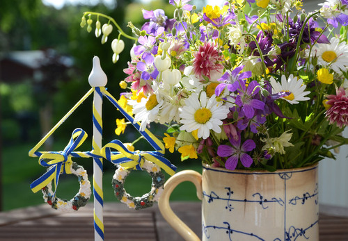 Närbild på bukett med sommarblommor i en kanna bredvid en midsommarstång i miniatyr.