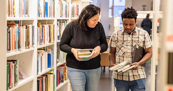 Två personer går mellan bibliotekshyllor och diskuterar böcker de håller i händerna.