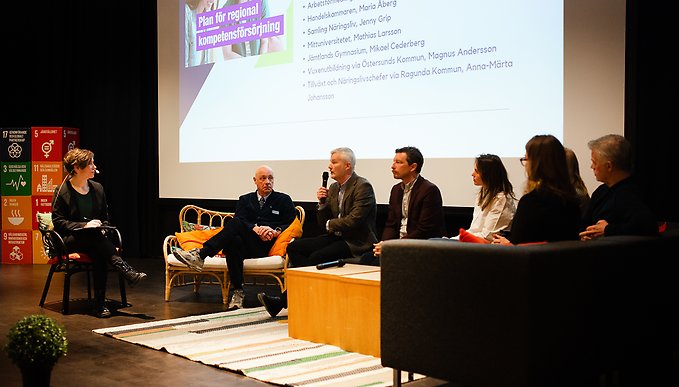 Flera talare sitter och samtalar i en soffa på scen, med en presentationsskärm bakom sig.